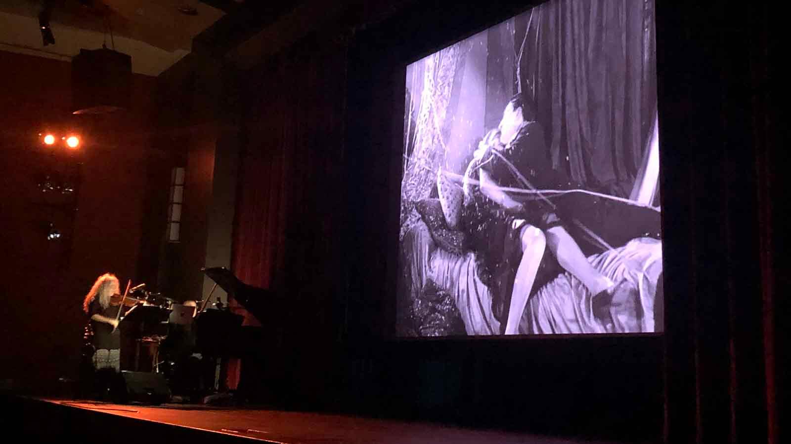 Violinist on lower left stage with film on large screen on right